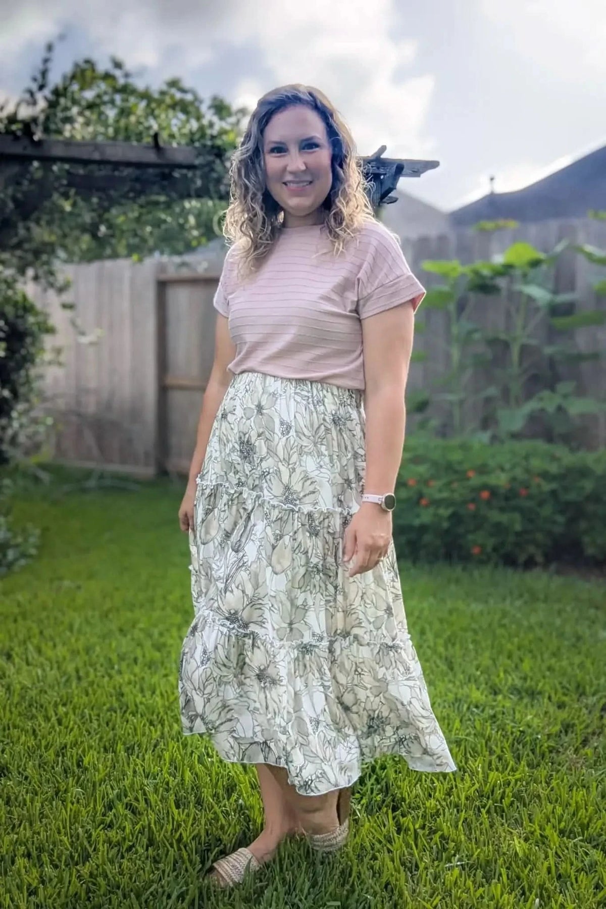 A woman wears a tiered floral skirt and a pink striped top in a grassy yard.