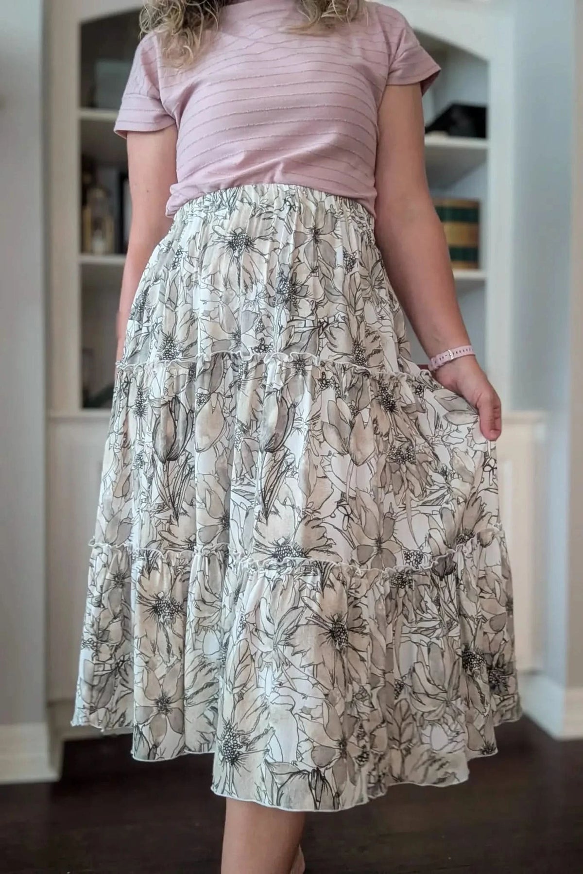A woman wears a tiered skirt with a sepia floral sketch pattern and a pink top.