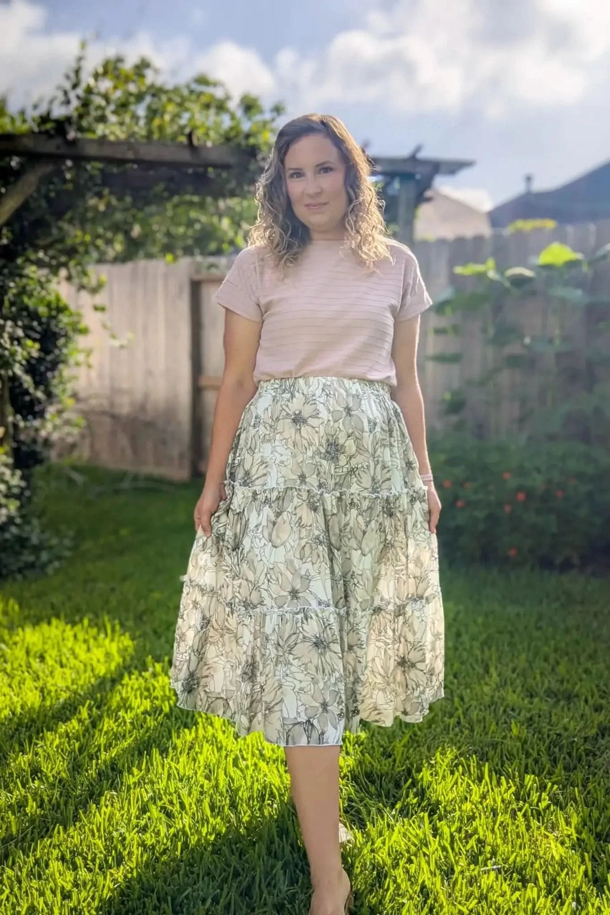 A woman in a sepia sketch floral skirt and light pink top stands in a sunny yard.