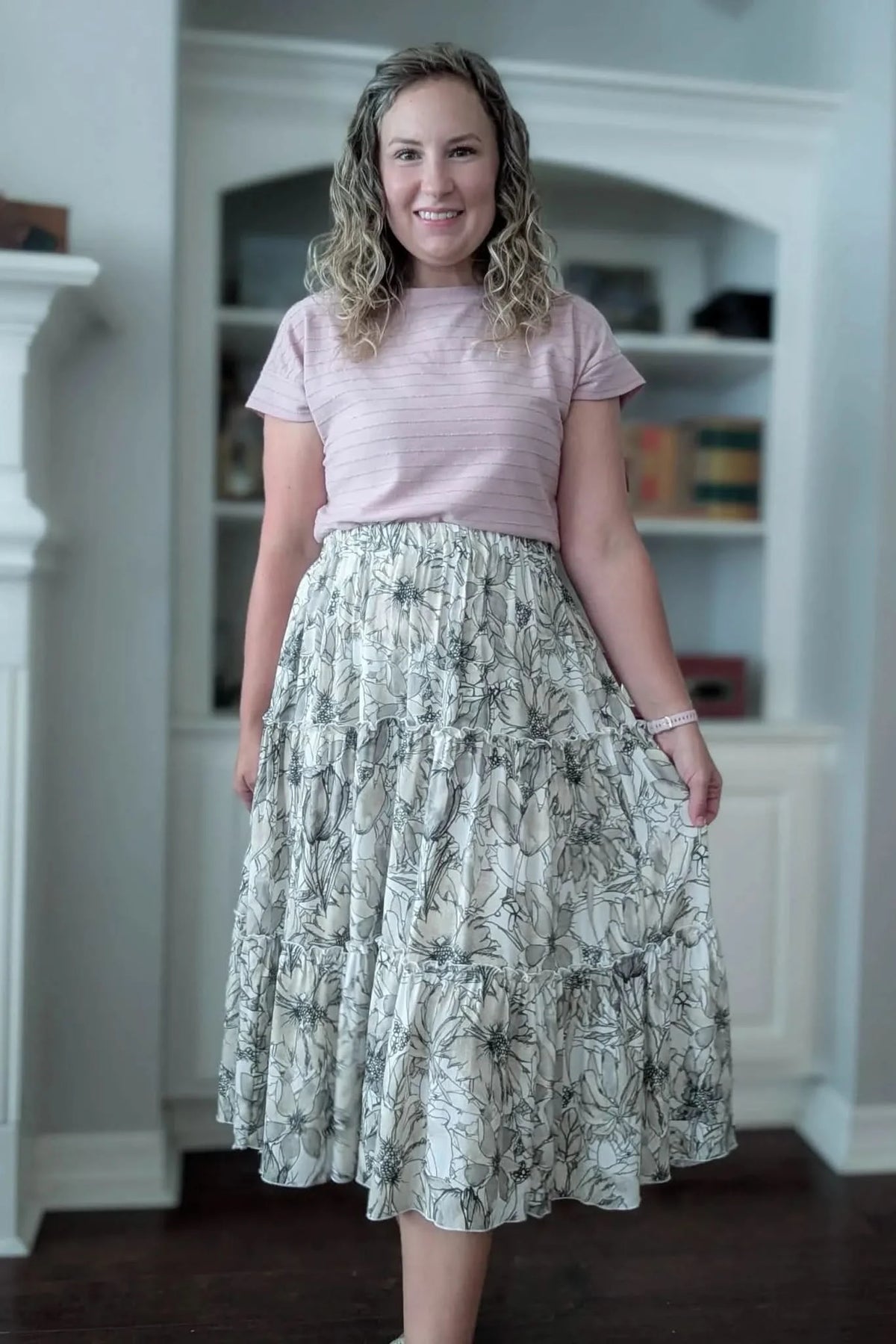 A woman smiles, wearing a pink top and a tiered skirt with a sepia floral sketch pattern.
