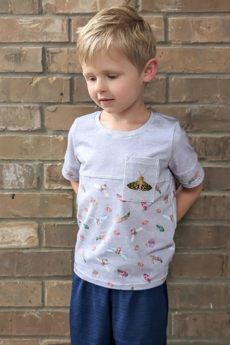 A young boy wears a light gray t-shirt with a colorful bug pattern and a moth on the pocket.