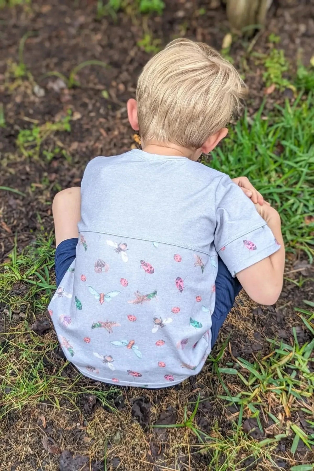 A child in a bug-patterned shirt kneels on dirt and grass.