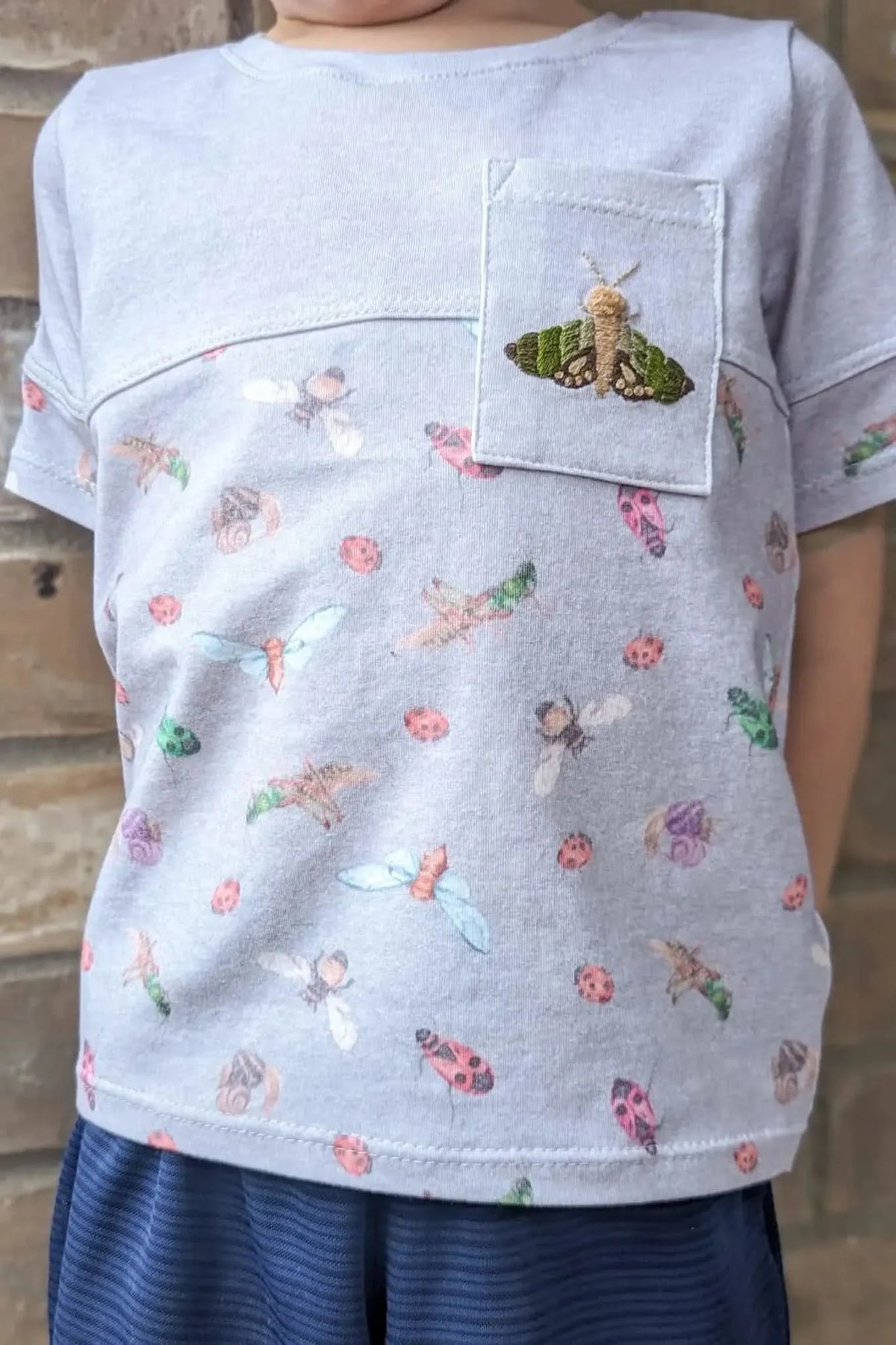 A child wears a light gray shirt with a colorful bug pattern and an embroidered moth.