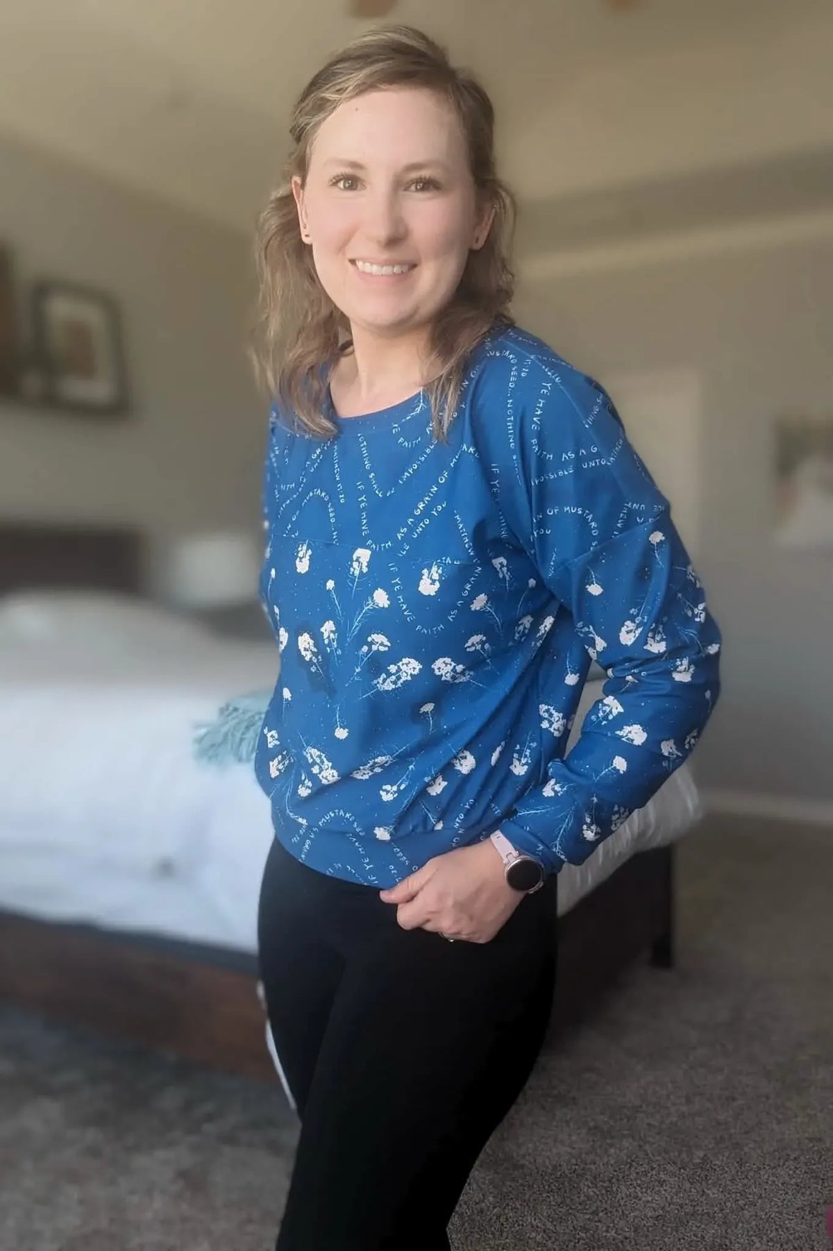 A woman smiles wearing a blue long-sleeved top with white floral and text patterns.