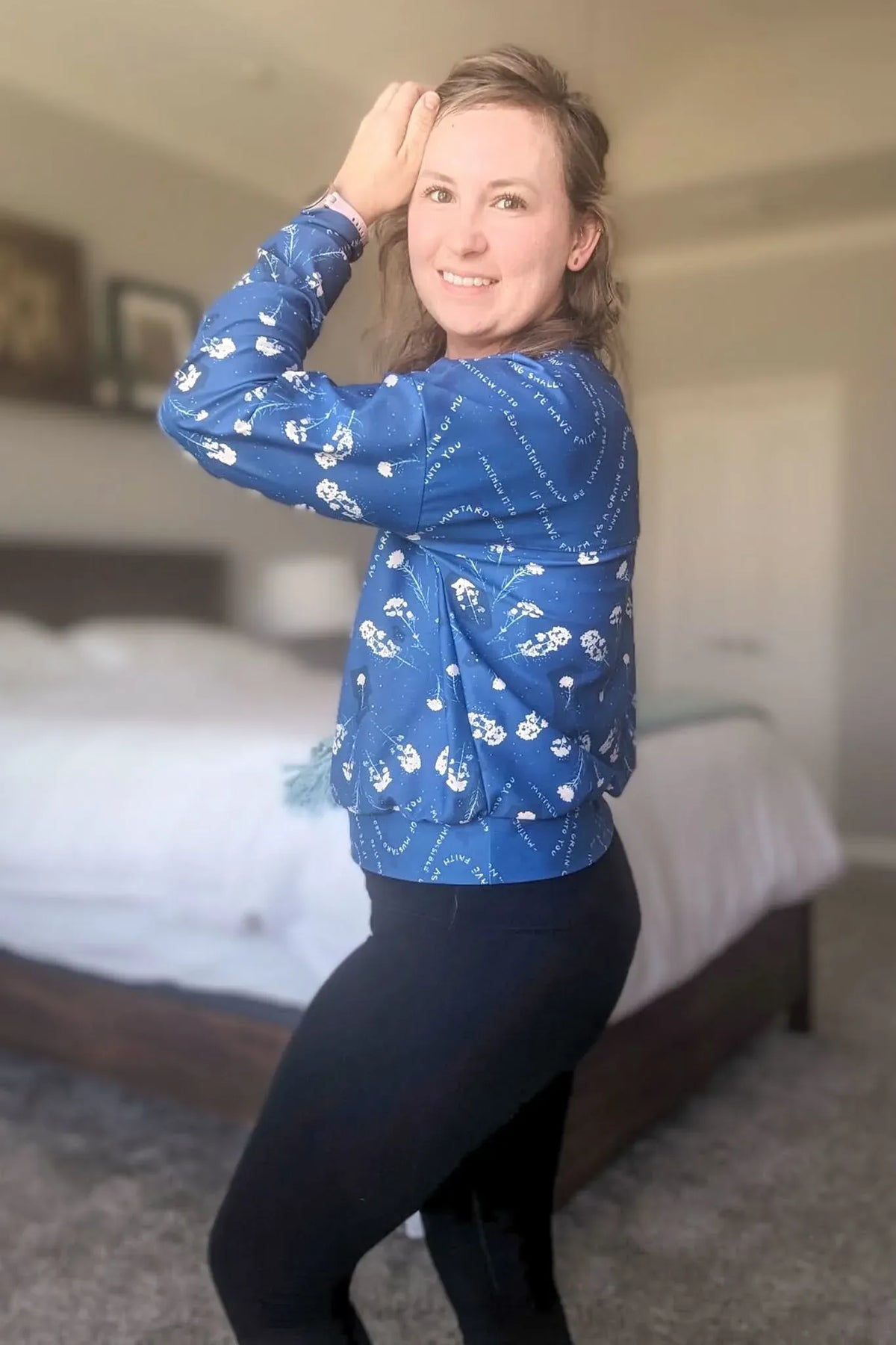 A woman in a blue mustard seed print top and black leggings smiles at the camera.