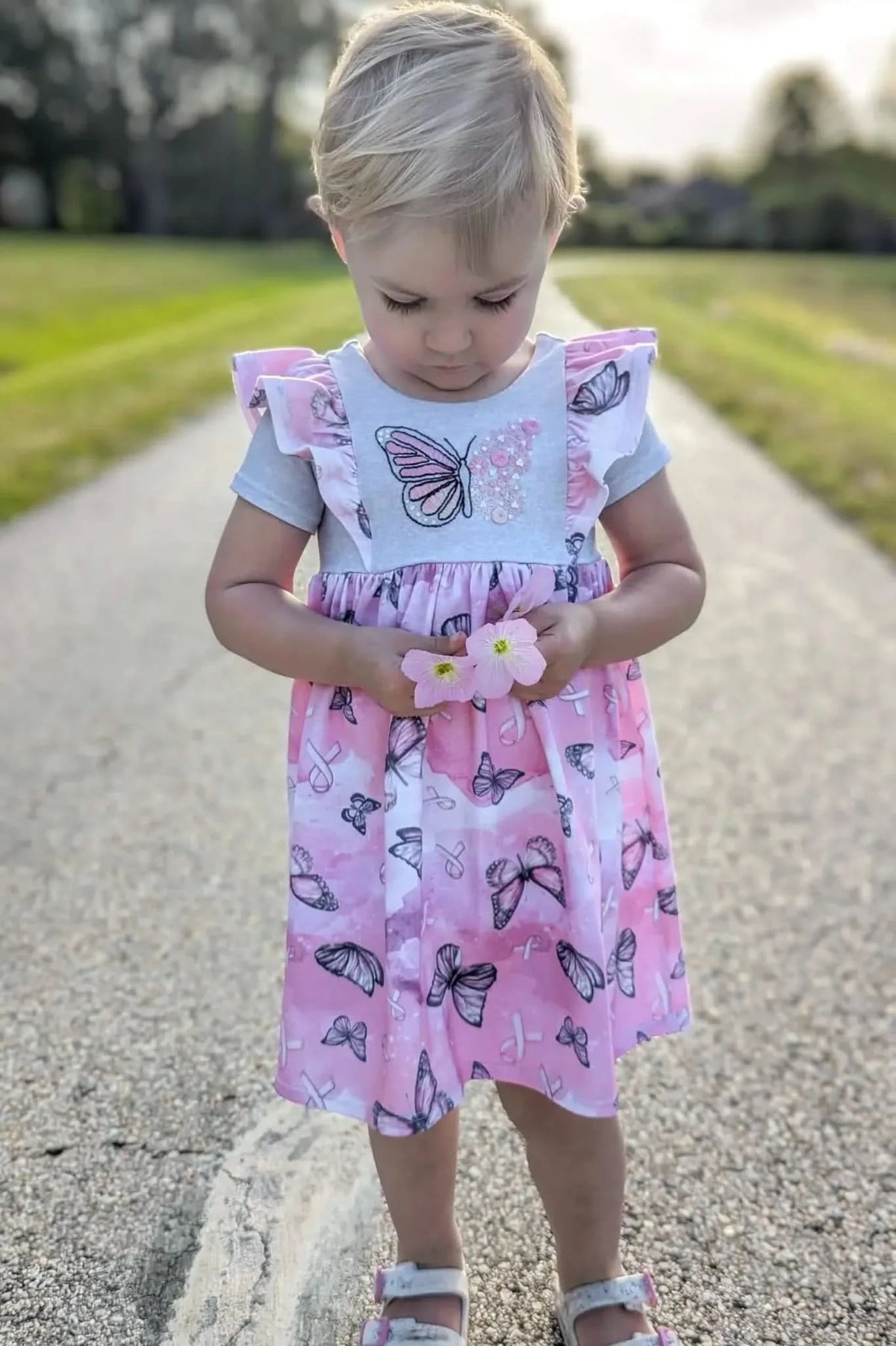 A young child in a pink butterfly dress holds two small pink flowers.