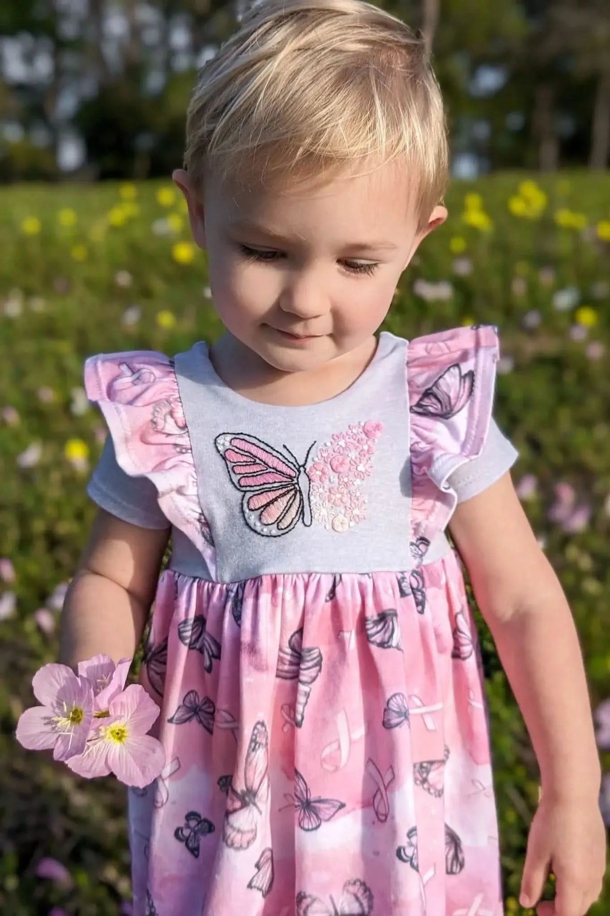 A young child in a pink butterfly dress holds pink flowers in a field.