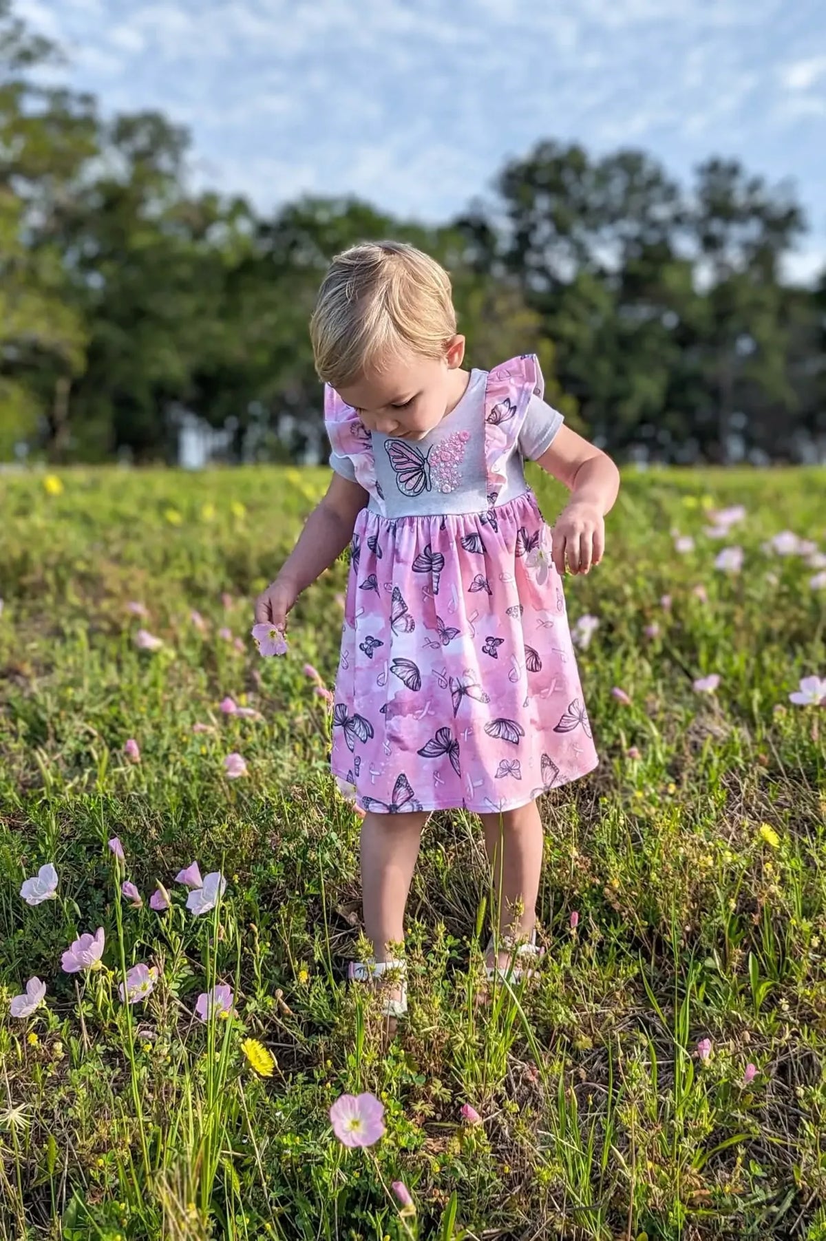 A young child in a pink butterfly dress picks a flower in a field.