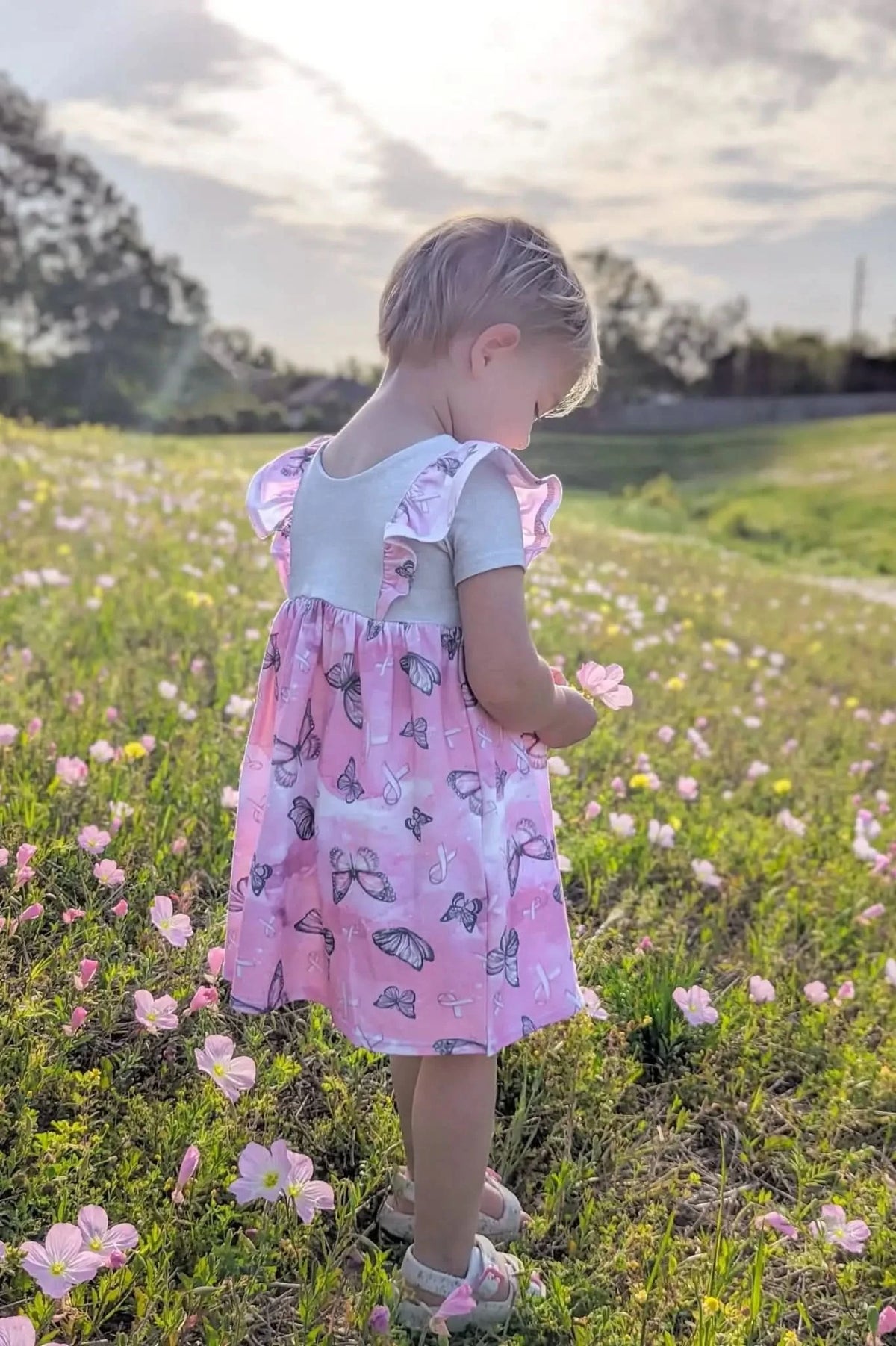 A young child in a pink butterfly dress holds a flower in a field.