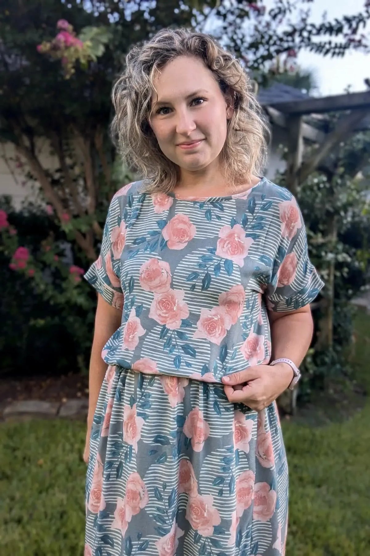 A woman in a floral and striped dress stands in a garden.