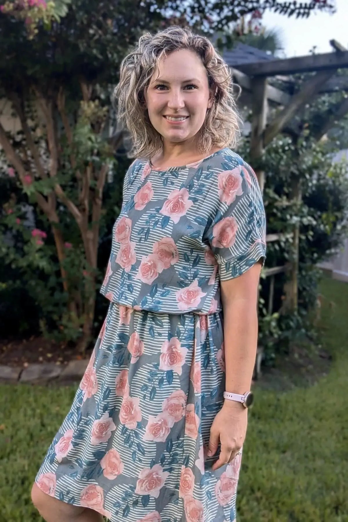 A woman in a floral and striped dress smiles in a garden setting.