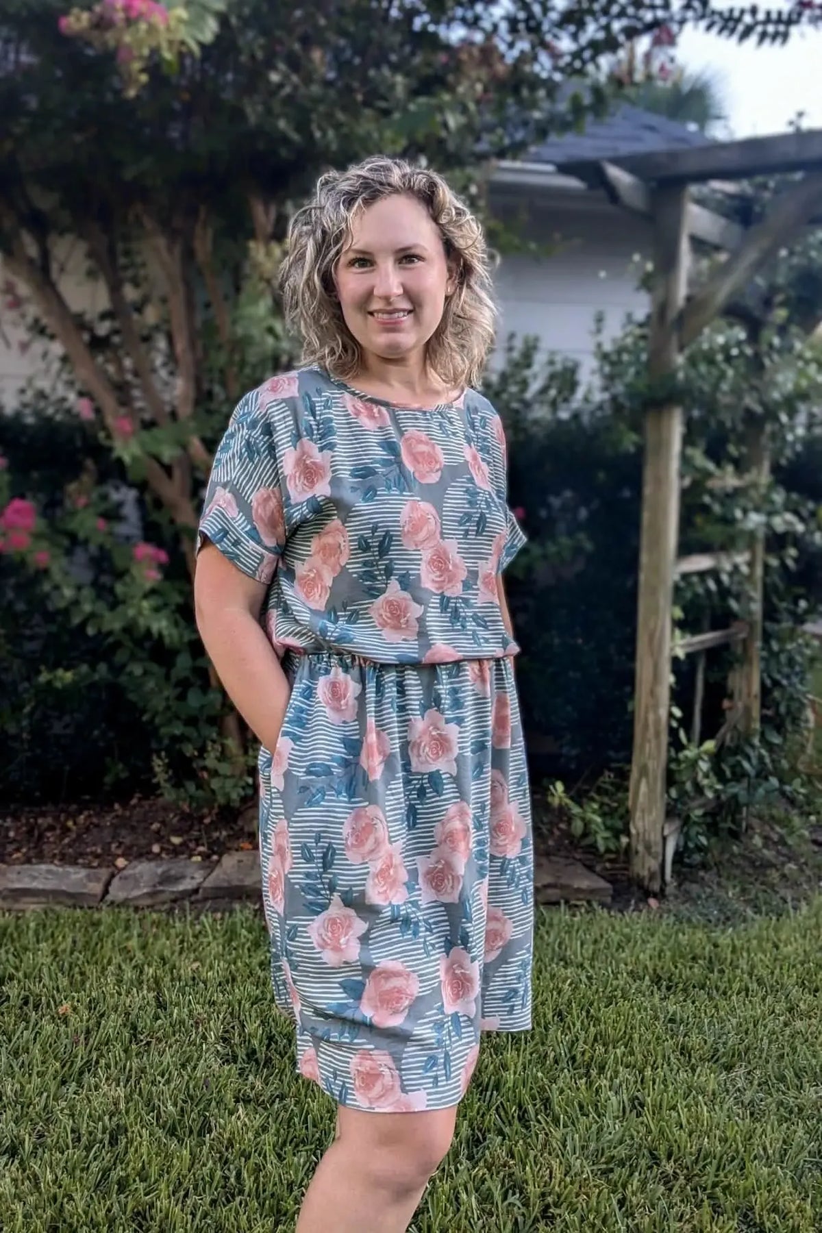 A woman in a floral and striped dress stands in a garden.