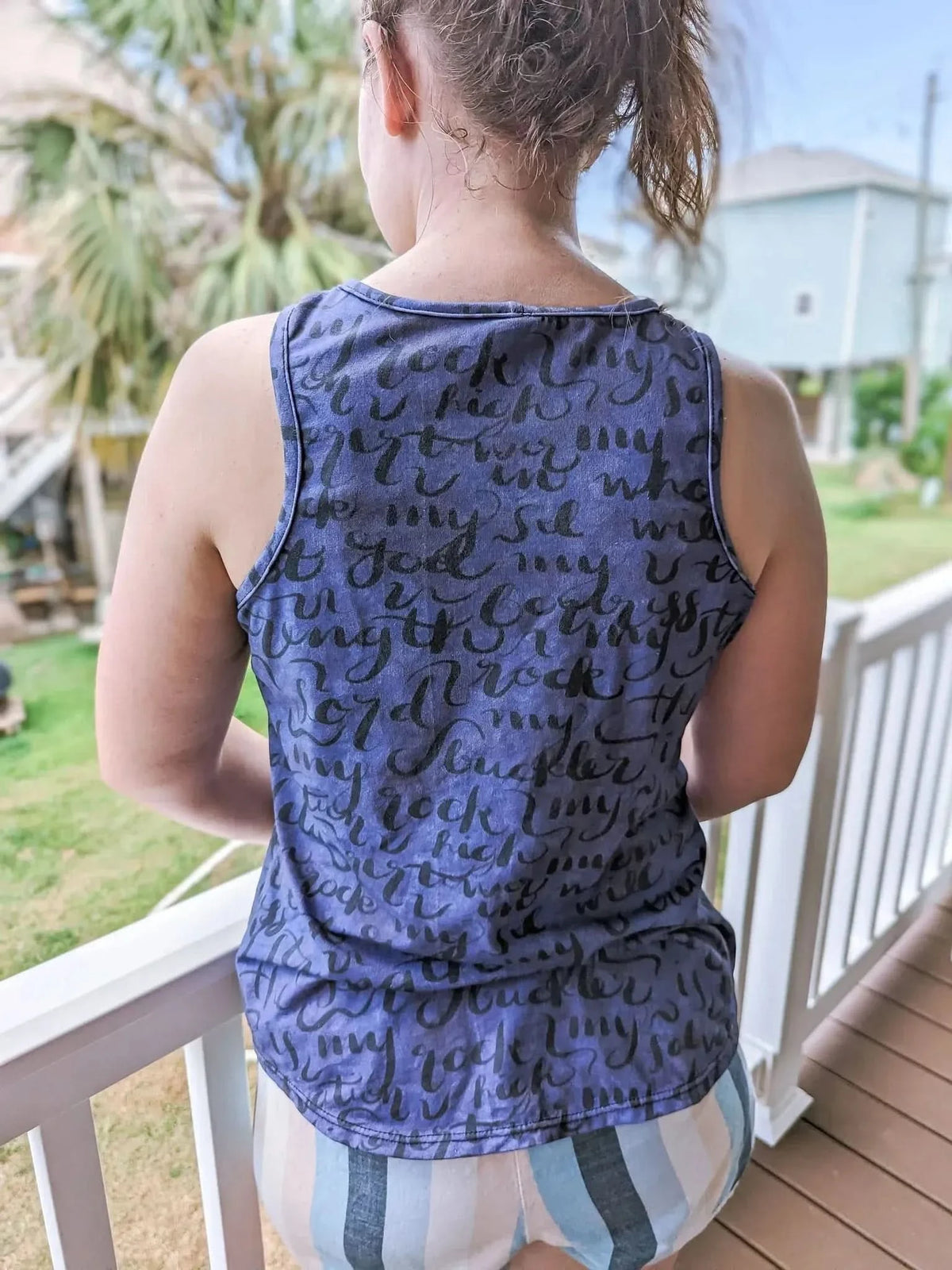 A woman from behind wears a blue tank top with black script and striped shorts.