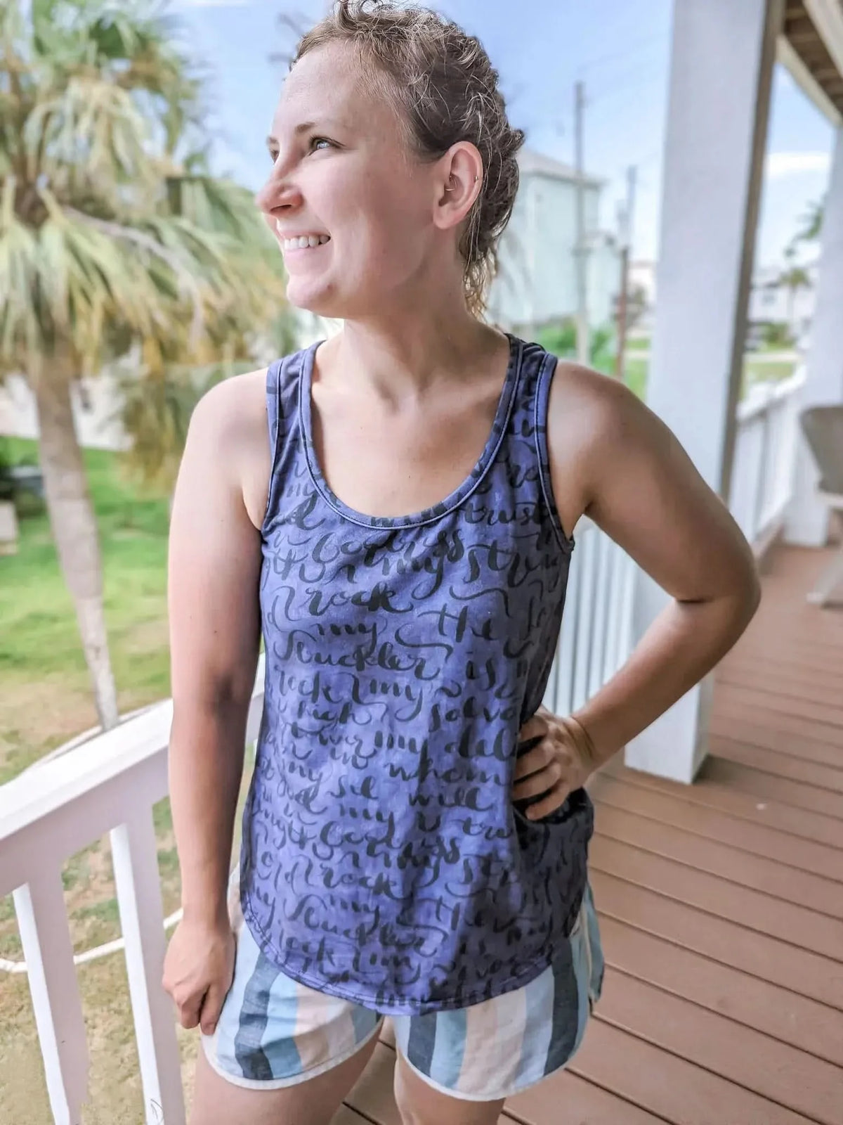 A woman smiles, wearing a blue tank top with black script and striped shorts.