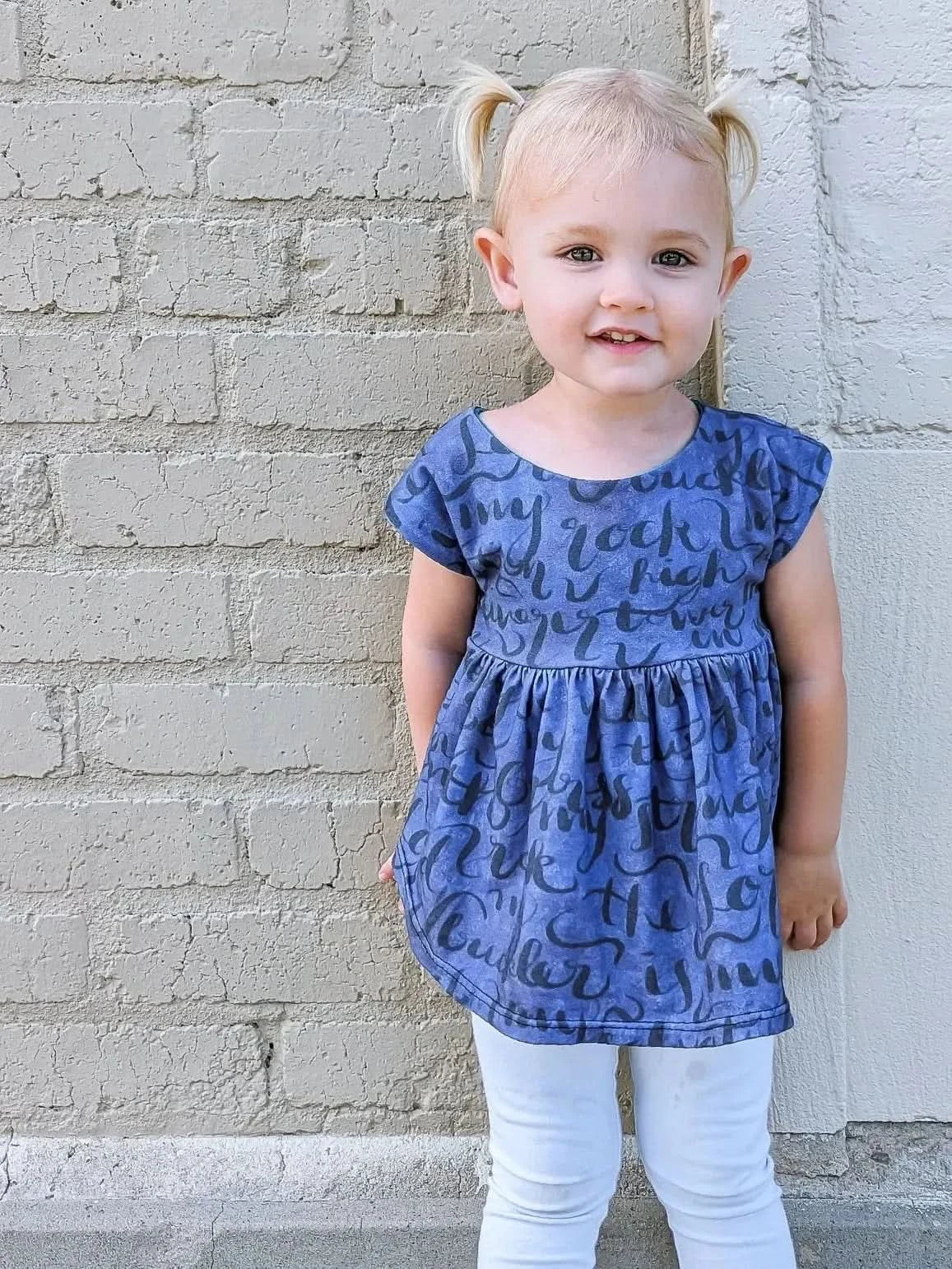 A young girl in a blue dress with "My Rock" text stands against a brick wall.