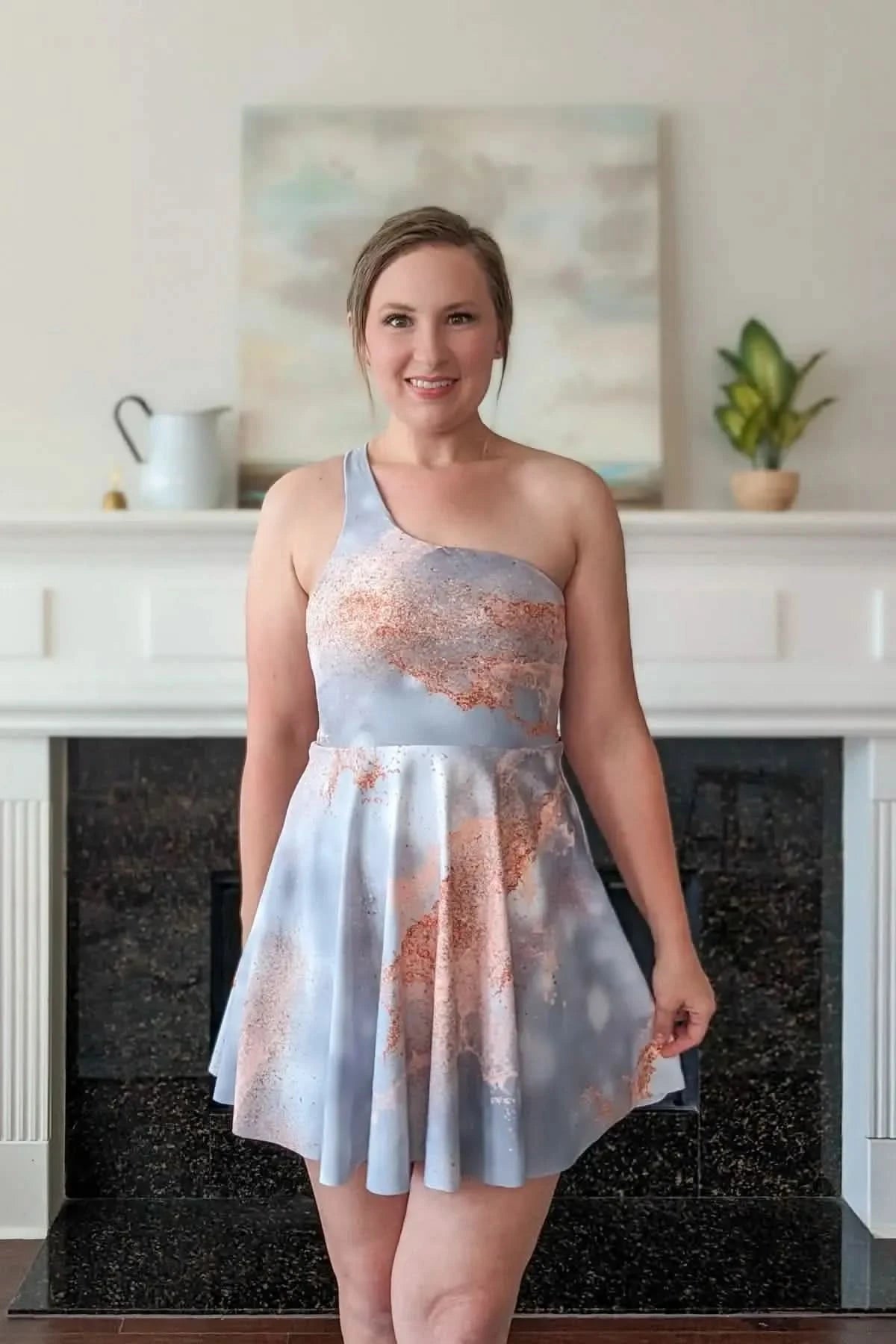 A woman wears a one-shoulder dress with a light blue and copper abstract pattern.