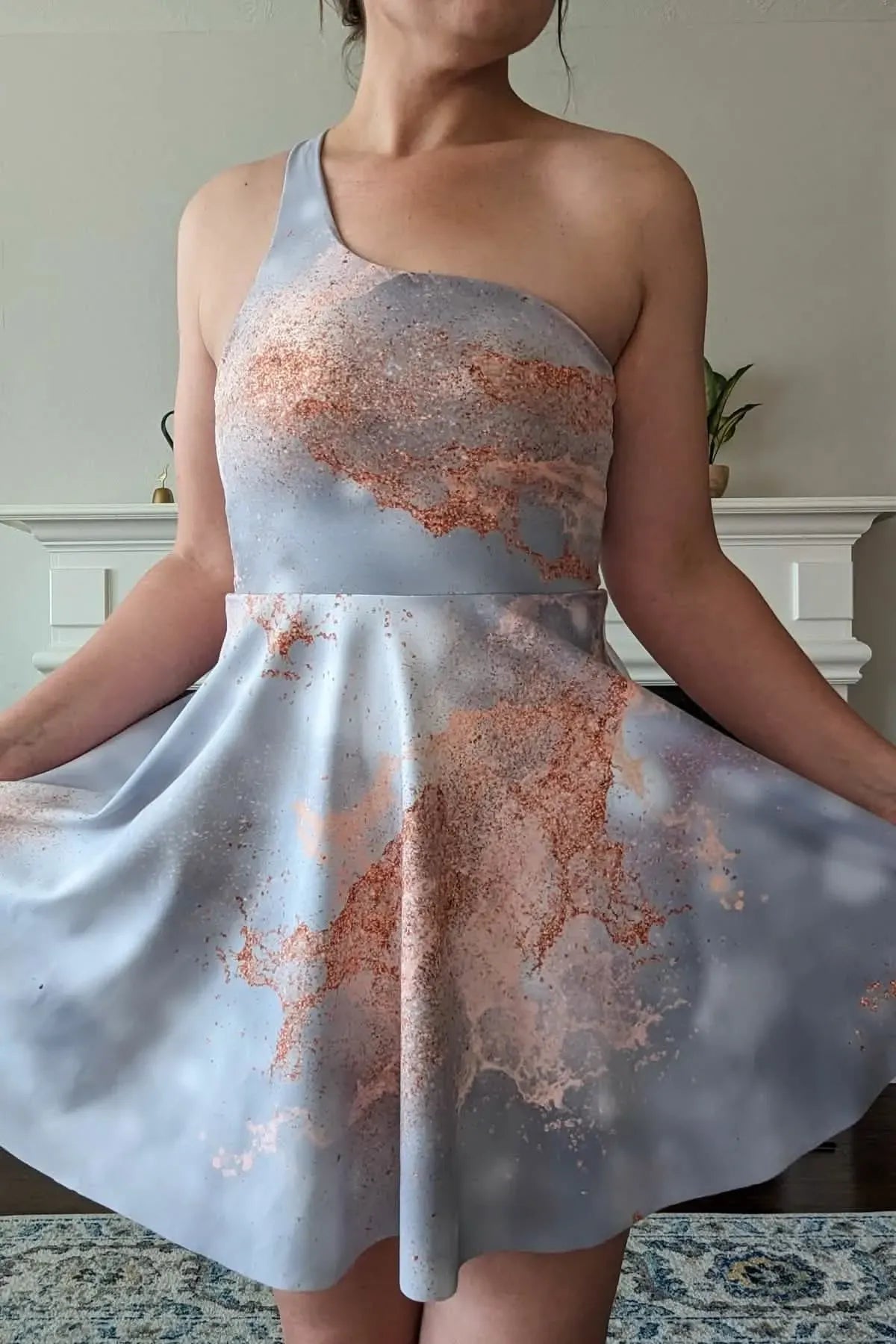 A woman models a one-shoulder dress with a gray and rose gold abstract pattern.