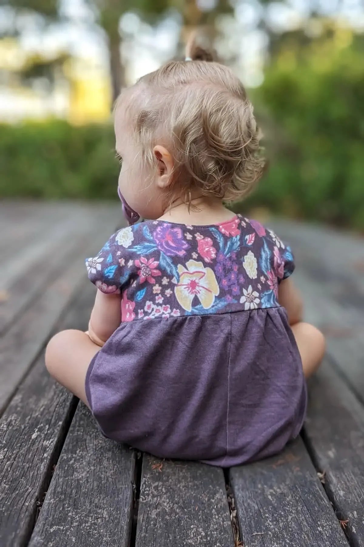 A baby in a maroon heather romper with a floral top sits on a wooden deck.