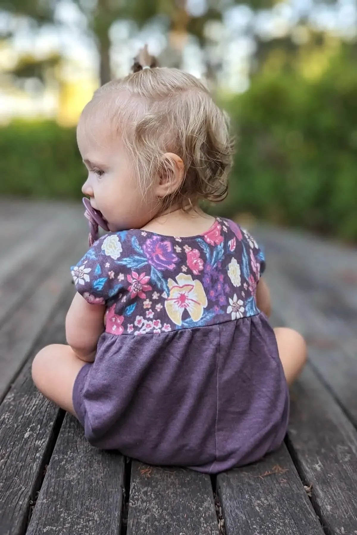 A baby wears a maroon heather romper with a floral top, sitting on a wooden deck.