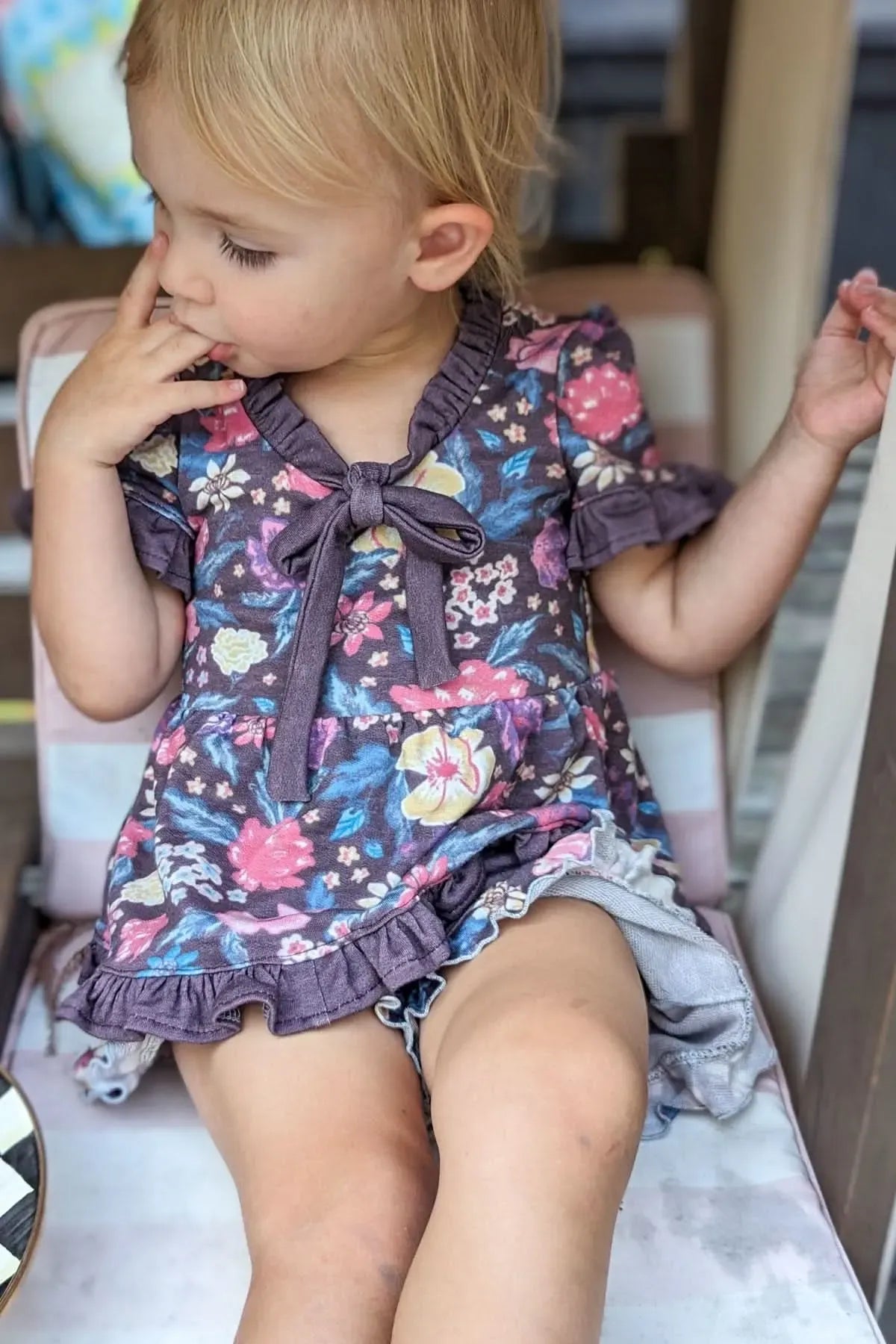 A young child wears a maroon floral dress with a bow, sitting on a striped cushion.
