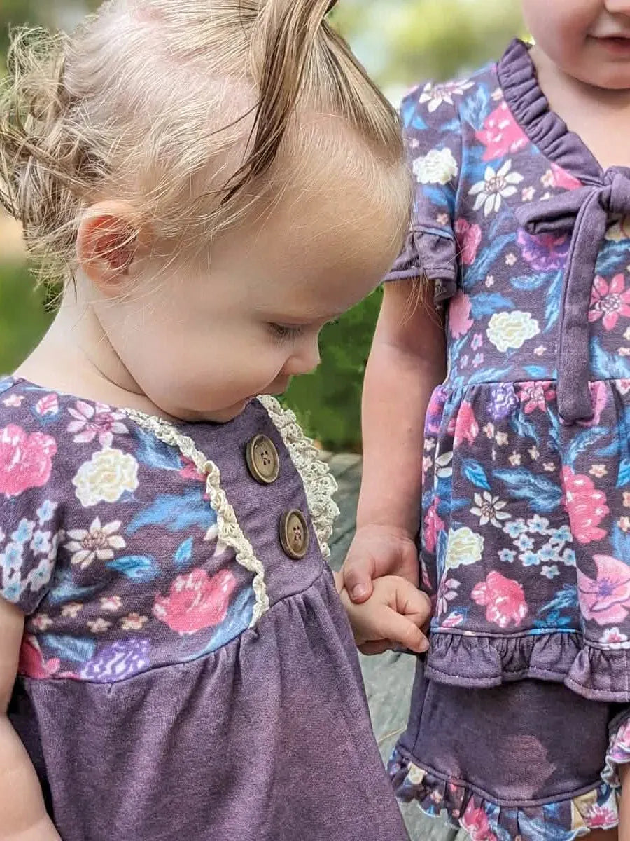 Two young children wear matching maroon floral outfits with ruffles and buttons.