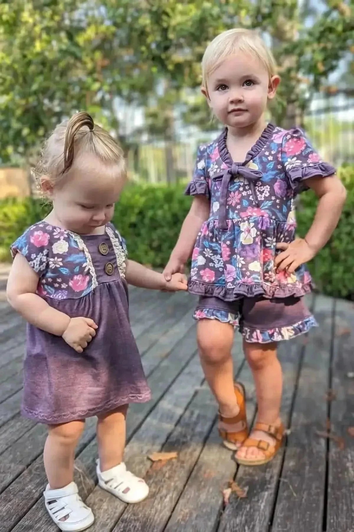 Two young girls wear matching maroon heather floral outfits on a wooden deck.