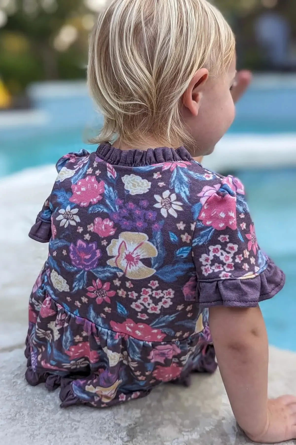 A child in a maroon floral dress with ruffled sleeves and collar sits by a pool.