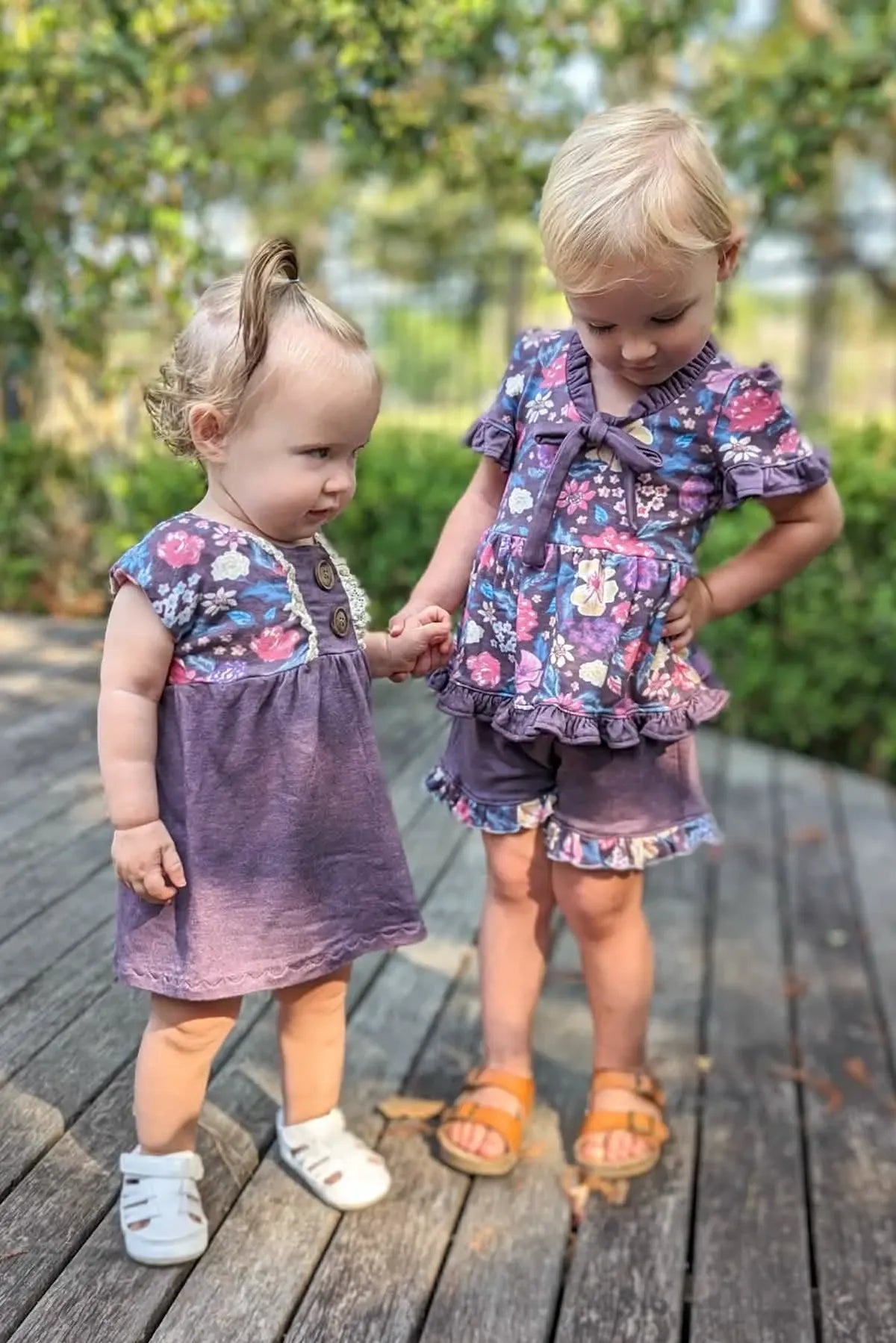Two young girls wear matching maroon heather floral outfits on a wooden deck.