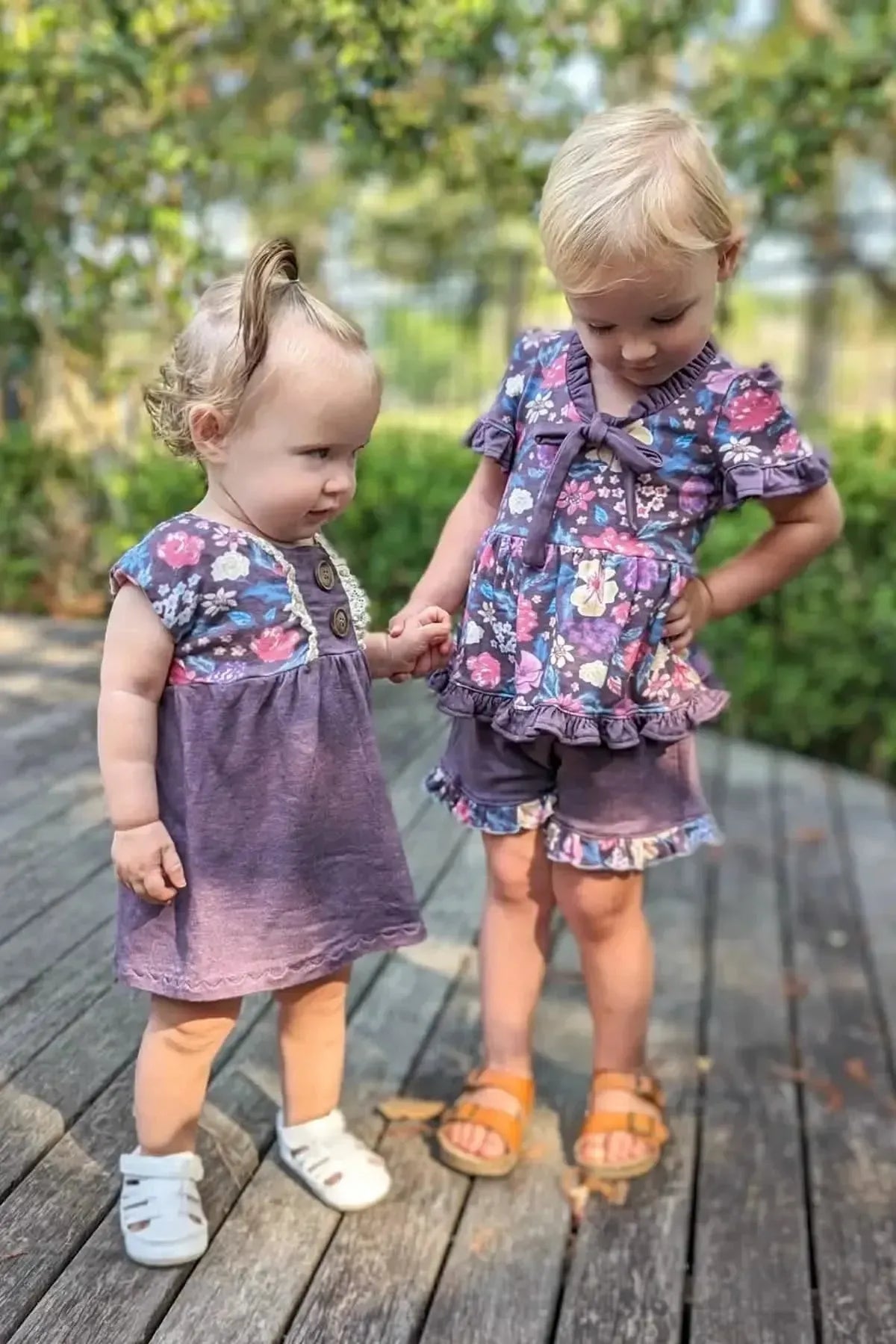 Two young girls hold hands outdoors, wearing matching maroon floral dresses with shorts.