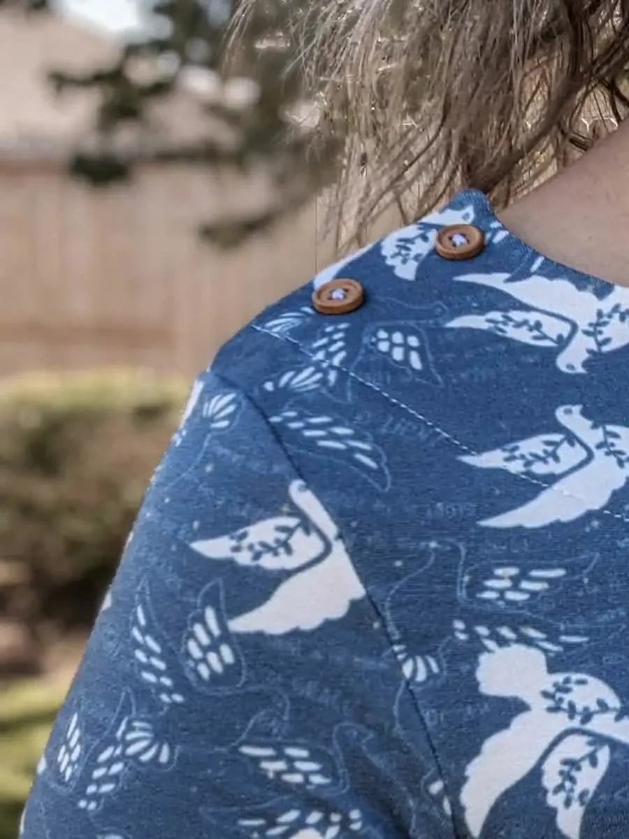 A person wears a blue top with white dove and leaf patterns, featuring two wooden buttons on the shoulder.