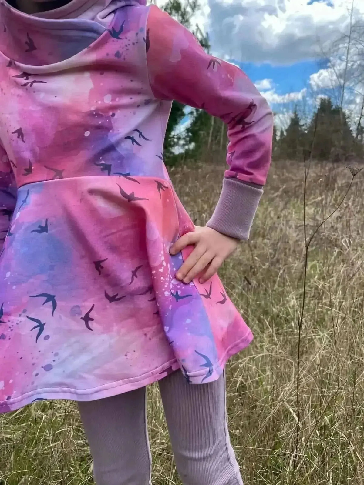 A child wears a pink and purple dress with a bird pattern in a field.