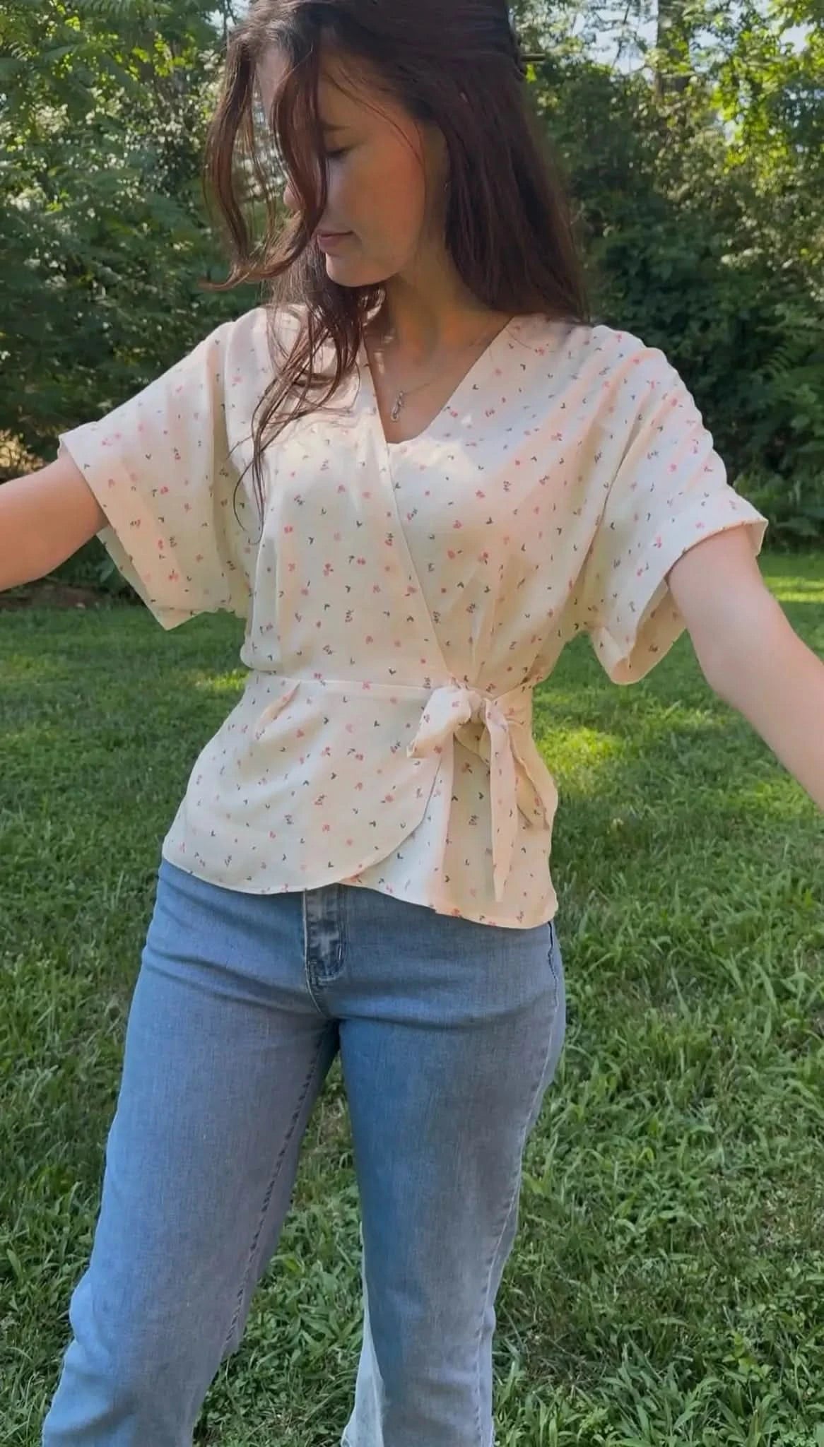 A woman wears a cream wrap top with small pink and green floral print and blue jeans.