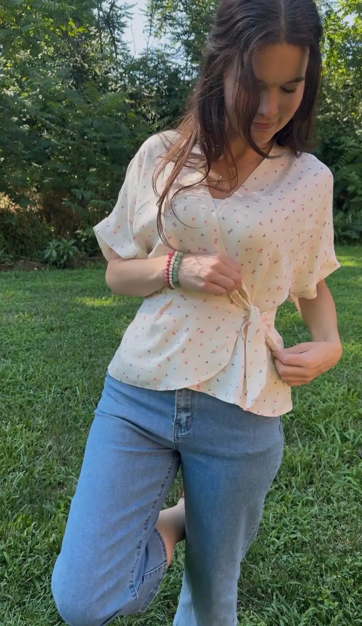 A woman in a floral wrap top and jeans kneels in a grassy outdoor setting.