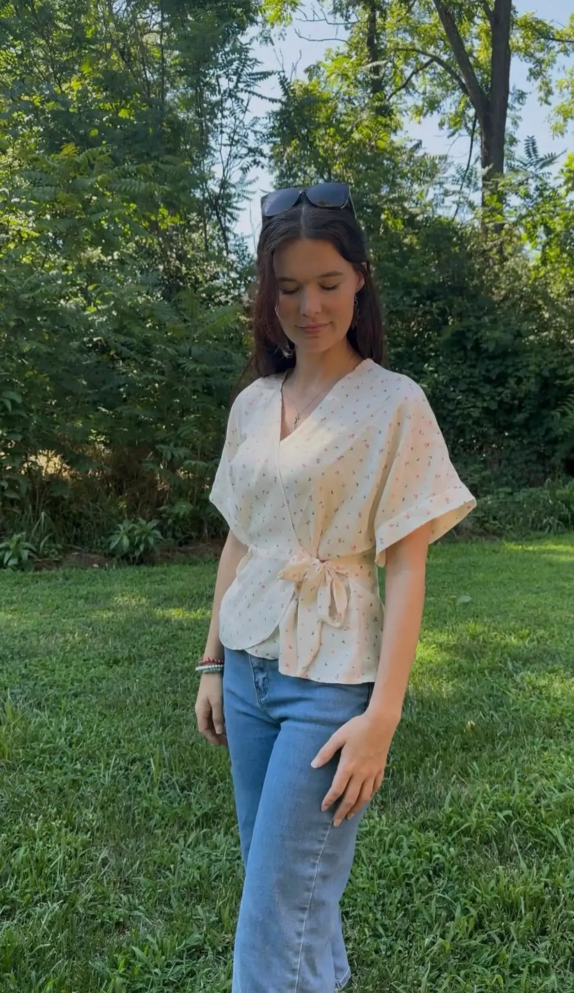 A woman wears a cream wrap top with small pink and blue floral patterns and blue jeans.