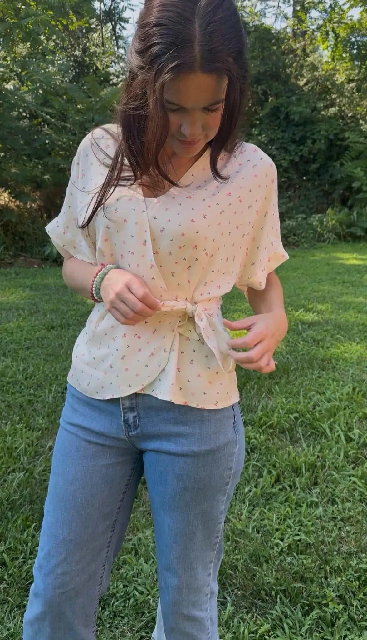 A woman in a floral wrap top and jeans ties the top in a grassy outdoor setting.