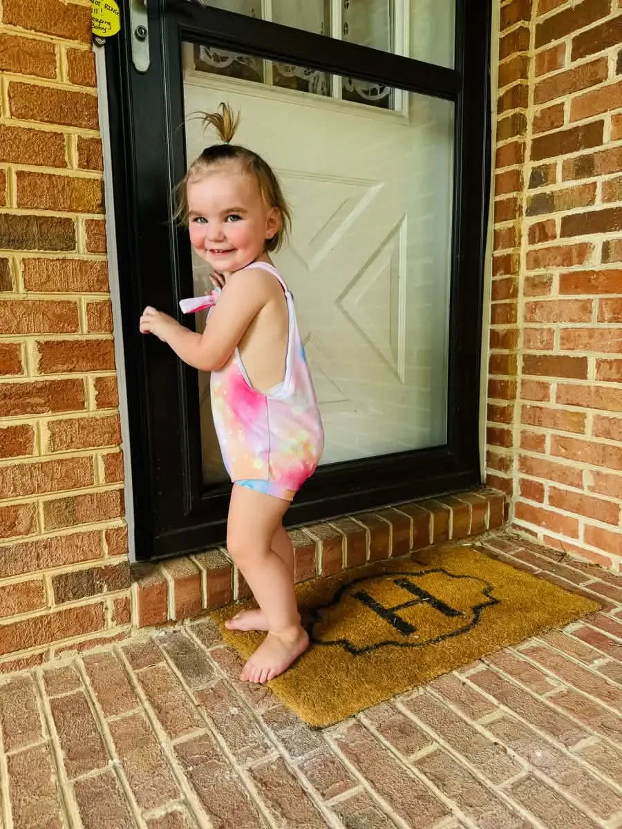 A smiling toddler wears a tie-dye romper with pink, purple, and blue hues.