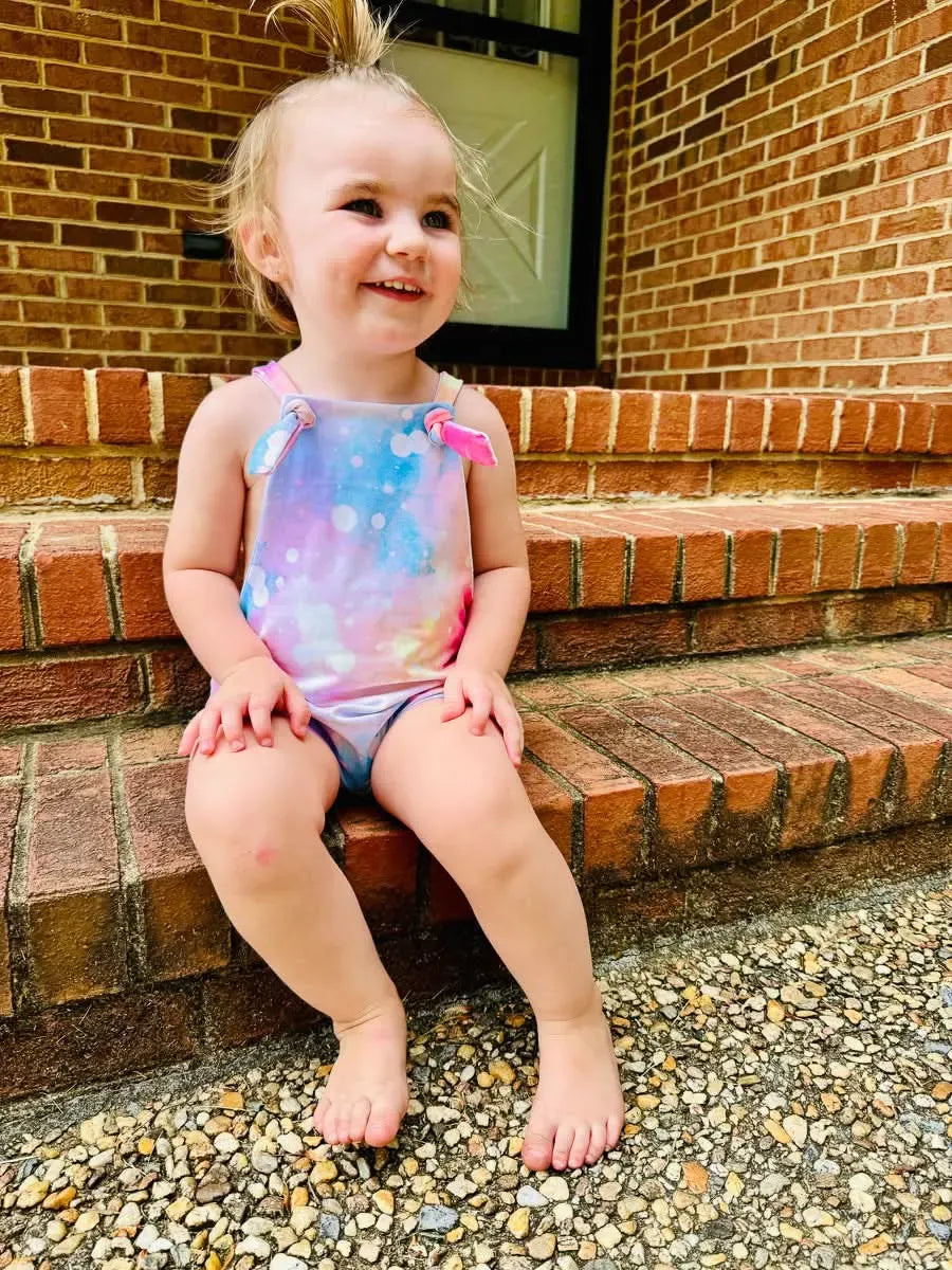 A smiling toddler wears a Cotton Candy Sparkle swimsuit while sitting on brick steps.