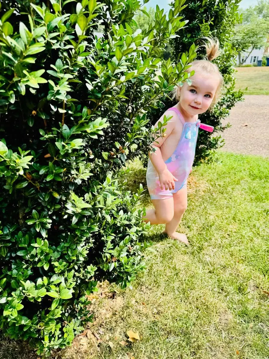 A young child in a cotton candy sparkle swimsuit stands next to a green bush.