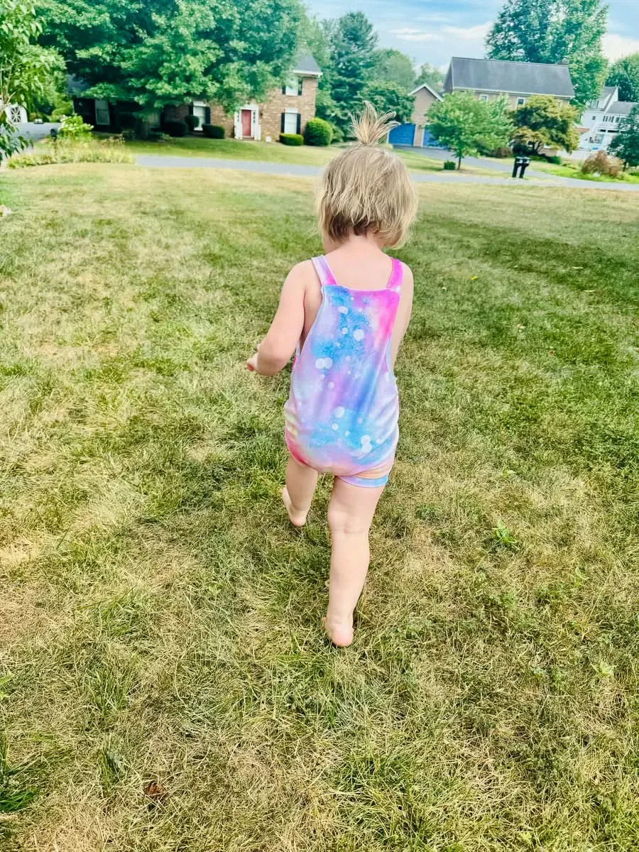 A child in a Cotton Candy Sparkle swimsuit walks away on a grassy lawn.