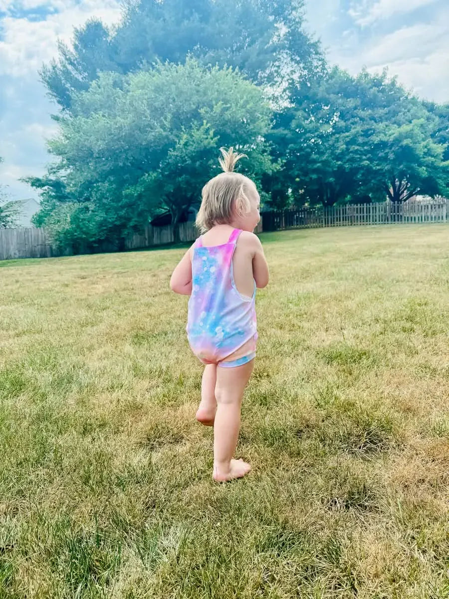 A toddler in a cotton candy sparkle swimsuit stands in a grassy yard.