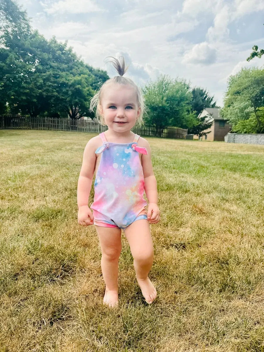 A young child wears a cotton candy sparkle romper with pink bow straps.
