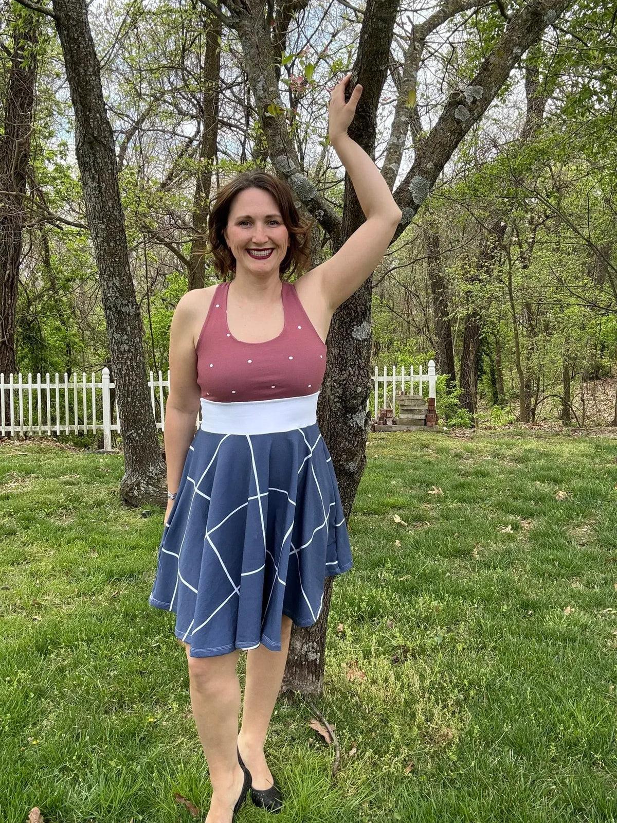 A woman in a navy windowpane skirt and a pink top stands by a tree.
