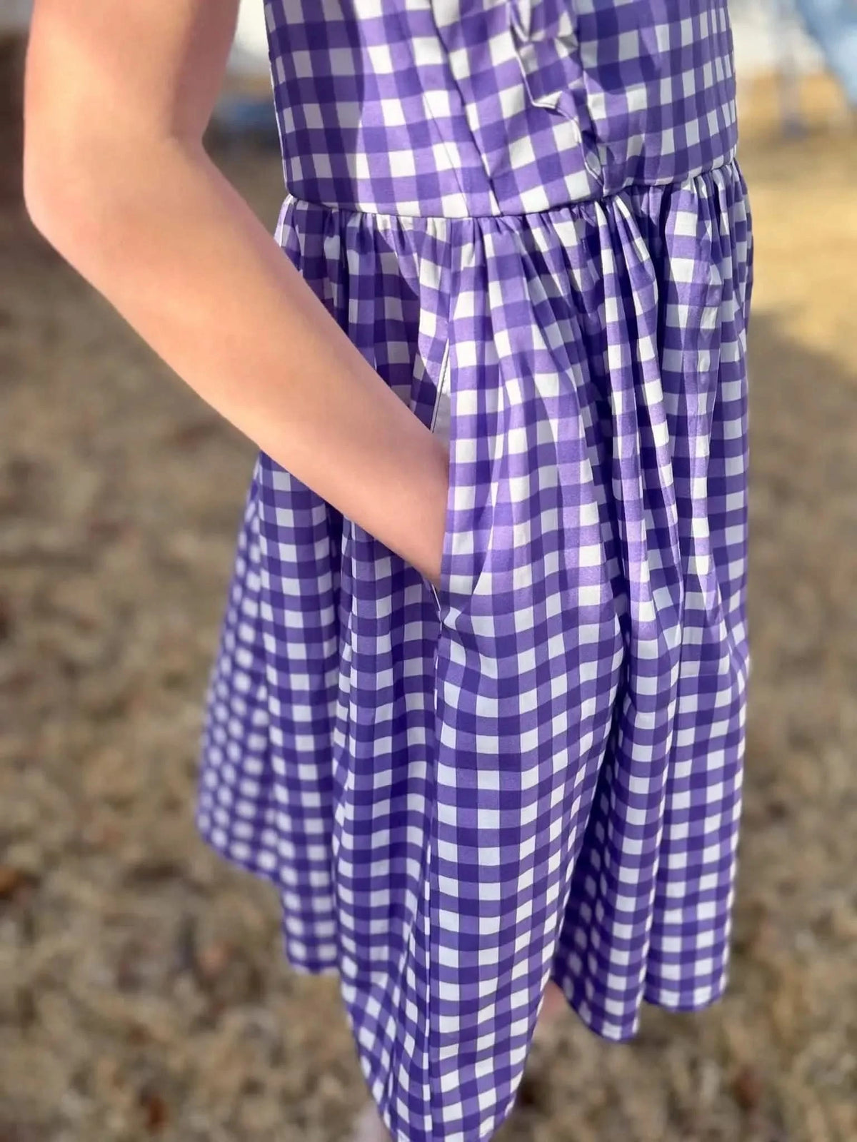 A person wearing a violet gingham dress with a hand in the pocket.