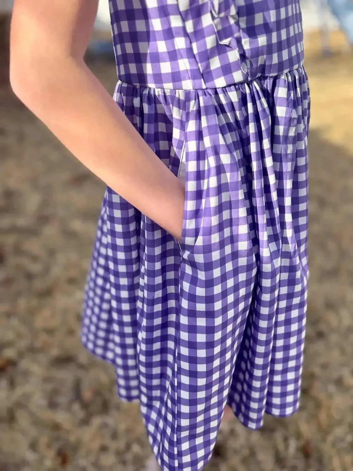 A person wears a violet and white gingham dress with a hand in the pocket.
