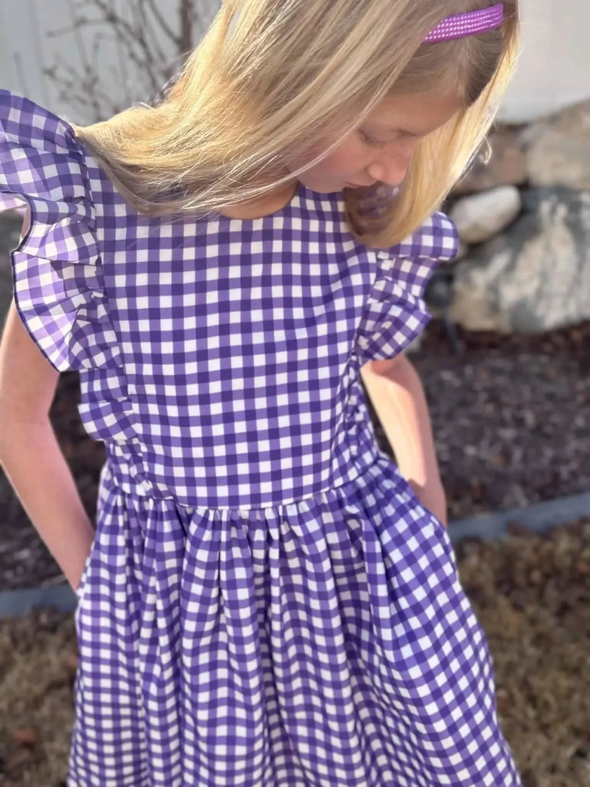 A young girl wears a violet gingham dress with ruffled sleeves and a purple headband.