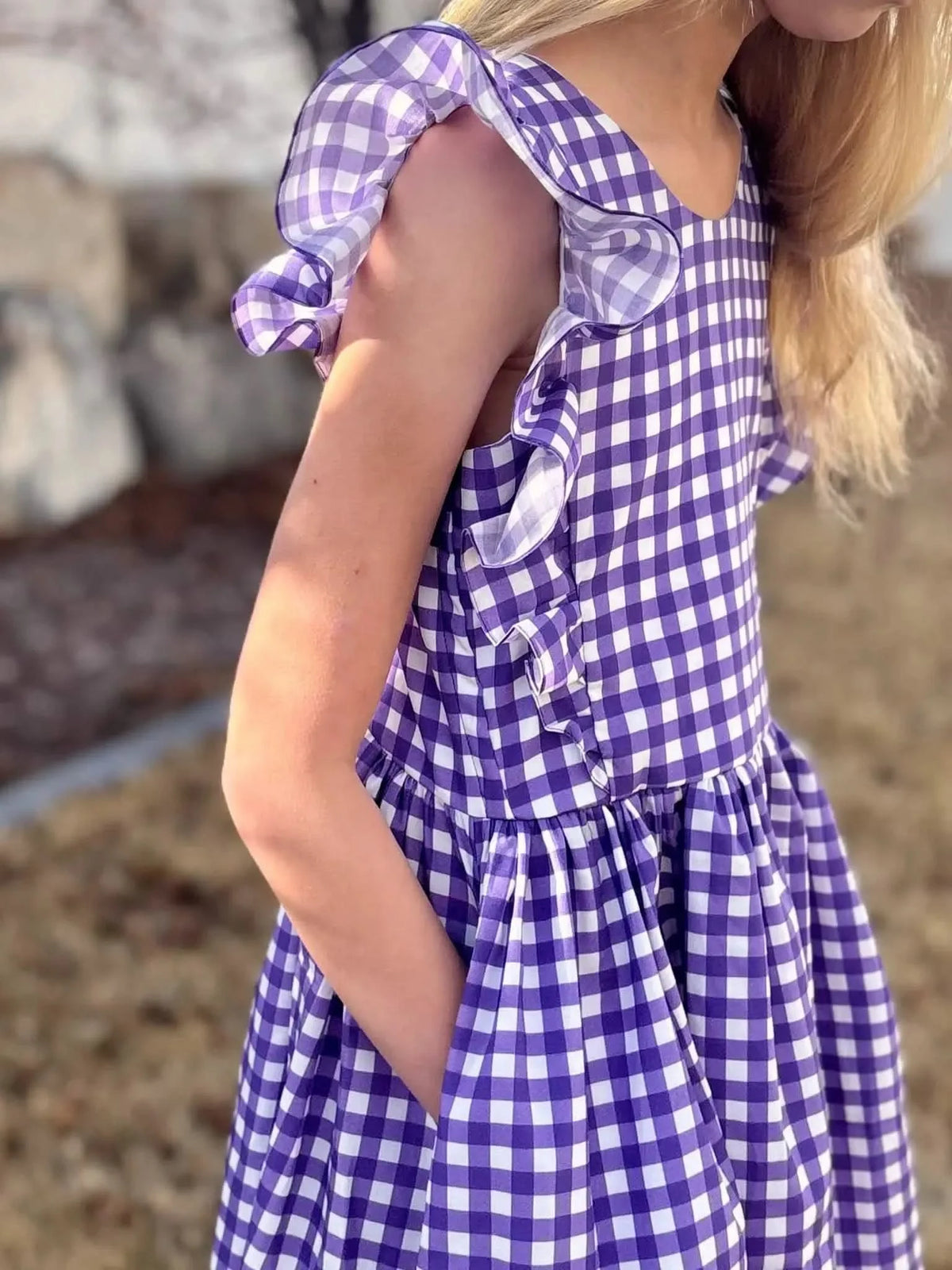 A child in a violet and white gingham dress with ruffled sleeves and pockets.