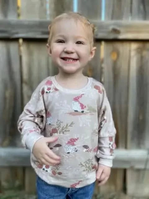A child wears a tan long-sleeved shirt with a gnome and mushroom pattern.