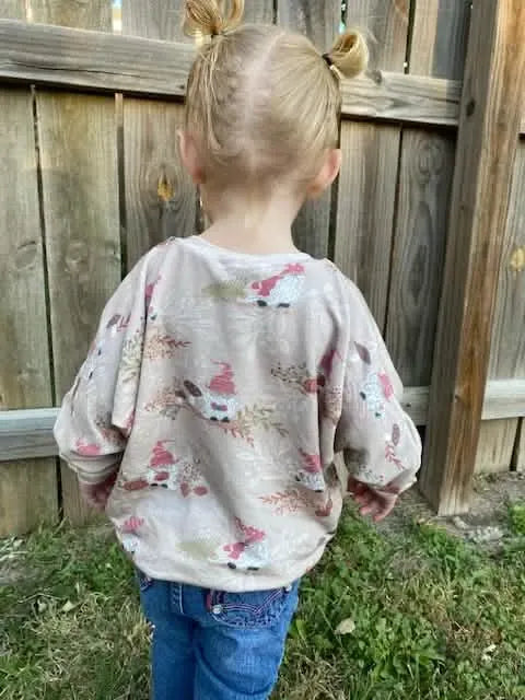 A child from behind wears a tan shirt with a gnome toile pattern and blue jeans.