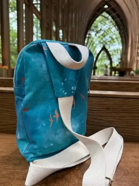 A dark teal bag with a sunset flight pattern and white straps sits on a wooden surface.