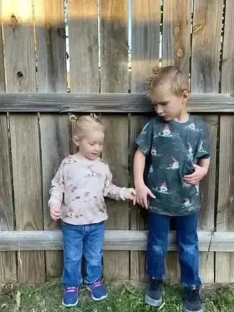 Two children wear shirts with a green gnome toile pattern, standing in front of a fence.